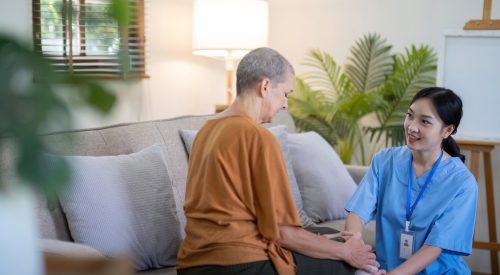 Asian caregiver kneeling beside elderly woman on couch at home. Concept of elderly care and support Asian caregiver kneeling beside elderly woman on couch at home. Concept of elderly care and support